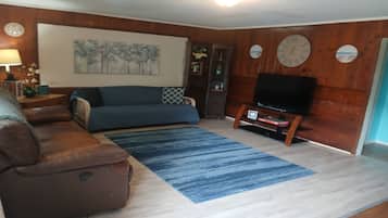 Un salon avec un canapé bleu, un fauteuil en cuir marron, un téléviseur à écran plat posé sur un meuble en bois, un tapis bleu et une horloge murale.