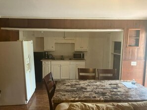 Eine kompakte Küche mit weißen Schränken, einer Mikrowelle und einem Kühlschrank. Es gibt einen Esstisch mit zwei Stühlen und ein Bett mit einem gemusterten Überwurf.