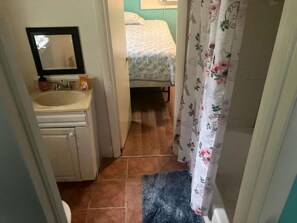 A narrow hallway with a bathroom on the left, a bed with a floral bedspread in the background, and a shower curtain with a floral pattern. - GPL Fort Myers Getaway (2670) (Fort Myers)