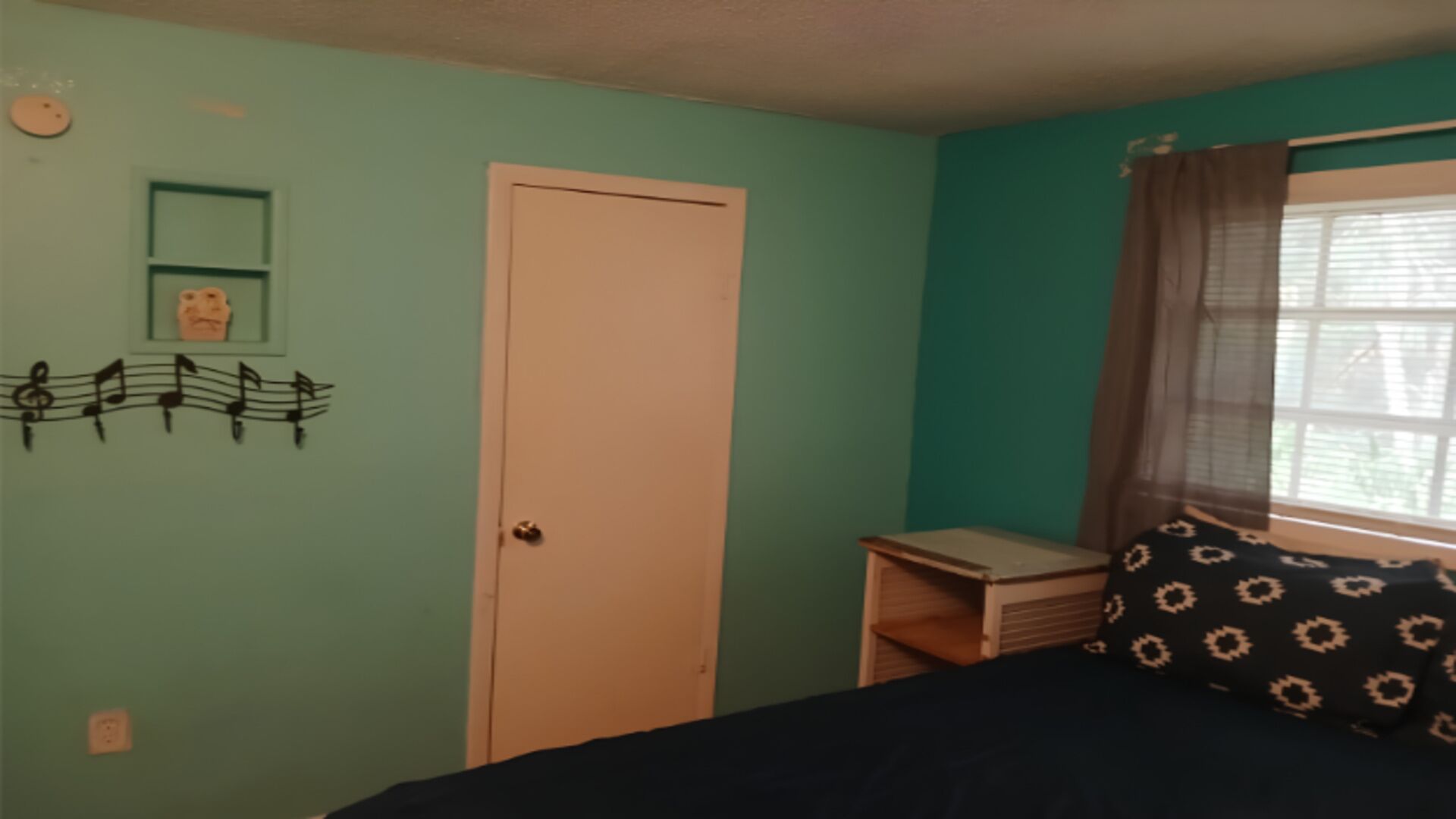 A room with teal walls, a closed door, a window with curtains, a bed with a patterned pillow, and a small table with a book.