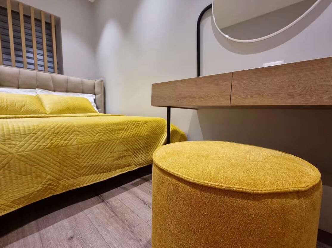 A bedroom with two beds, a wooden headboard, a round ottoman, and a mirror on the wall.