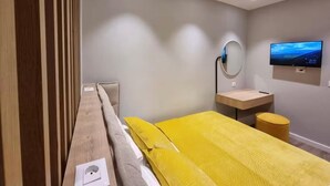 Une chambre d’hôtel moderne équipée d’un lit, d’un bureau, d’une chaise, d’un miroir rond, d’une télévision et d’un petit tabouret.