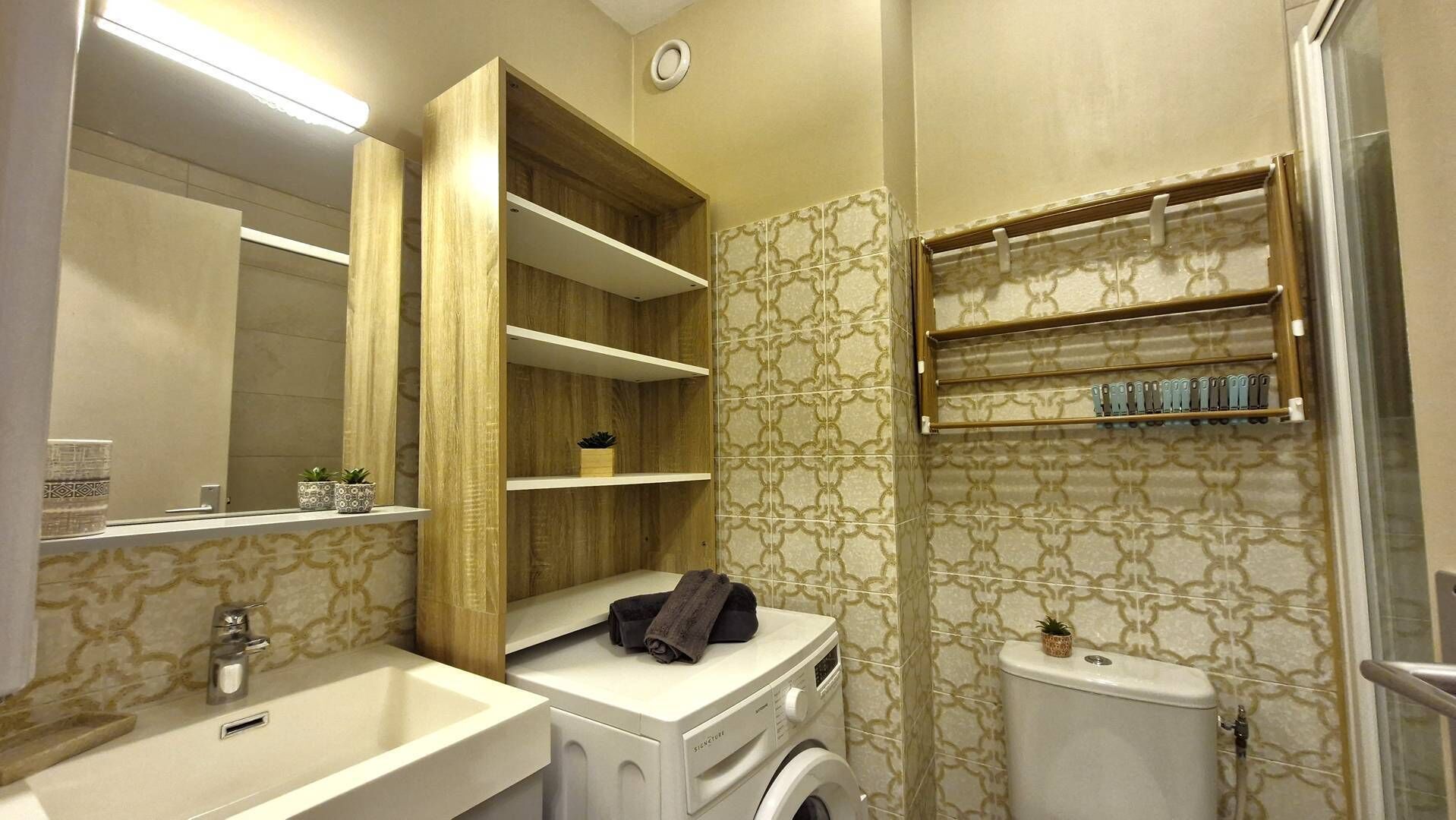 Un baño con lavabo, espejo, estantes de madera, lavadora e inodoro.