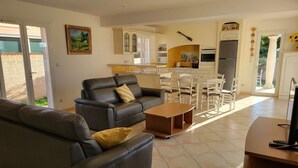 Ampla sala de estar com sofá, mesa de centro e mesa de jantar. Uma cozinha com armários brancos e painel de azulejos é visível ao fundo.