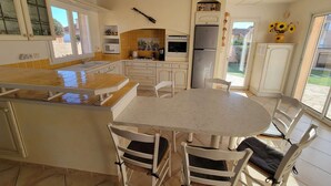 Cozinha com ilha central, armários brancos, micro-ondas, fogão e geladeira. Há uma área de refeições com uma mesa redonda e cadeiras.