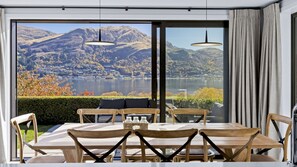 A dining area with a table and chairs, large windows with a mountain and lake view, and two pendant lights. - Perfect on the Peninsula with Lake View and Hot Tub (Kawarau Falls)