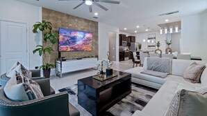 Ein modernes Wohnzimmer mit Fernseher, Deckenventilator und einer Küchenzeile im Hintergrund.