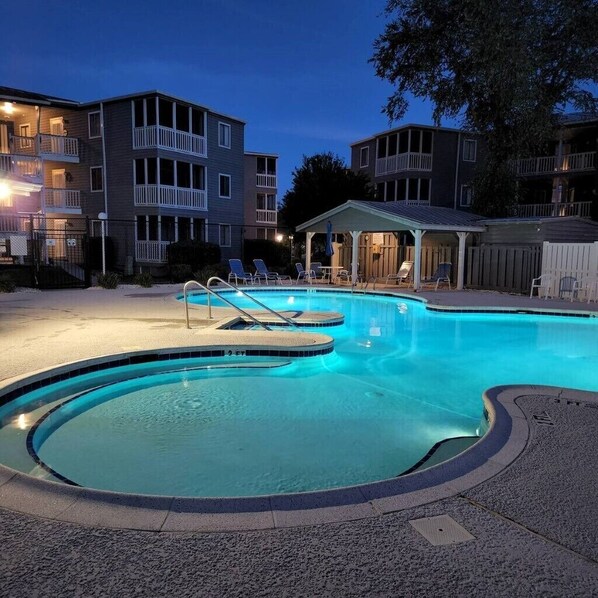 Una piscina con solárium, rodeada de edificios con balcones y una zona cubierta con sillas.
