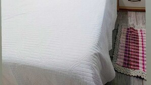 Un lit bien fait, avec une literie blanche, deux oreillers et des serviettes enroulées. On y trouve une table de chevet en bois, dotée de tiroirs, et une petite plante en pot.
