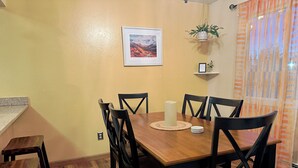 Un comedor con mesa de madera, sillas negras, un cuadro en la pared, planta en maceta y una ventana con cortinas.