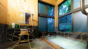 A stone-floored spa area with a wooden tub, a wooden shelf with bottles, and a window with a forest view.