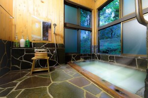 A stone-floored spa area with a wooden tub, a wooden shelf with bottles, and a window with a forest view.
