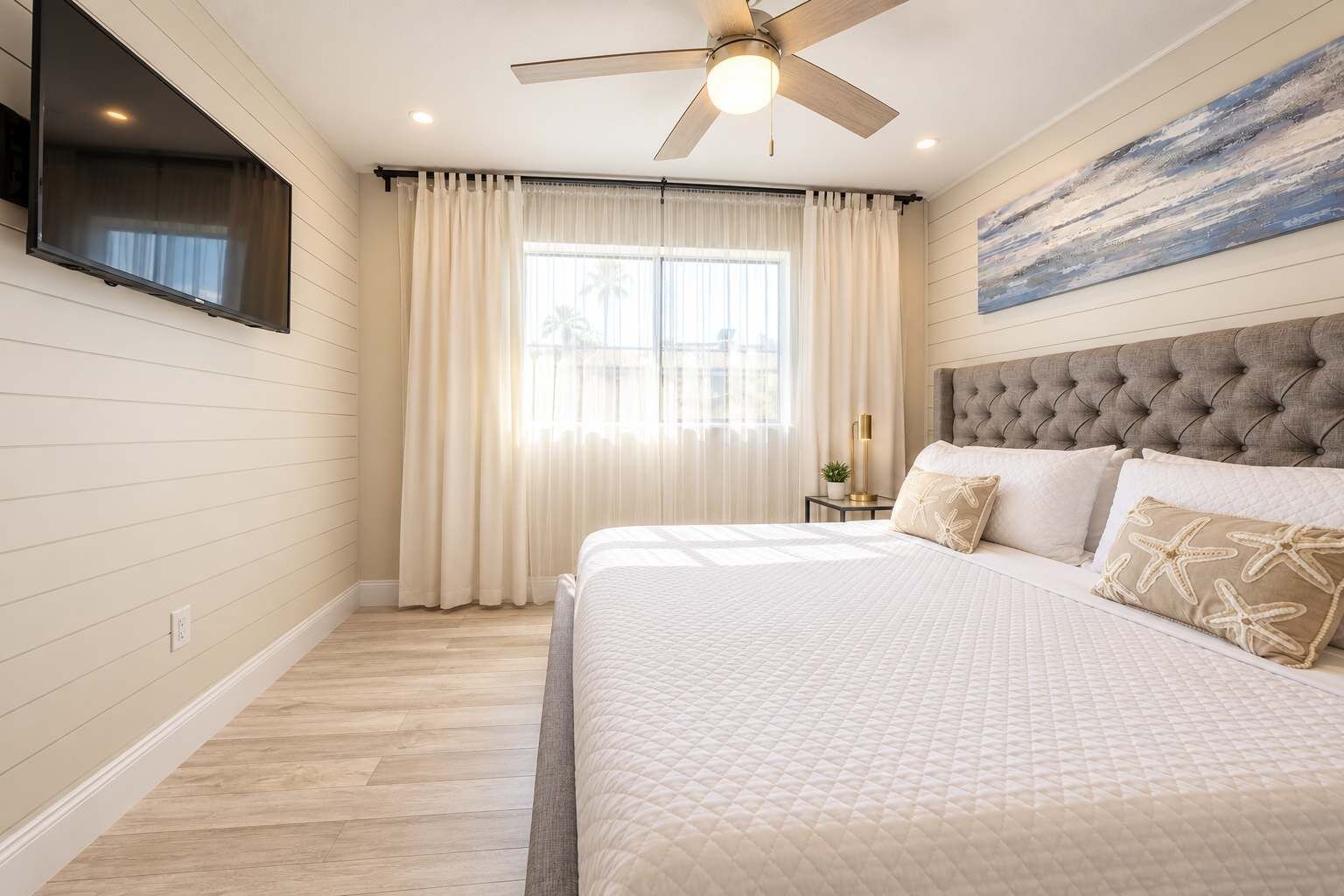 Un dormitorio con cama, ventilador de techo, televisor montado en la pared y ventana con cortinas.