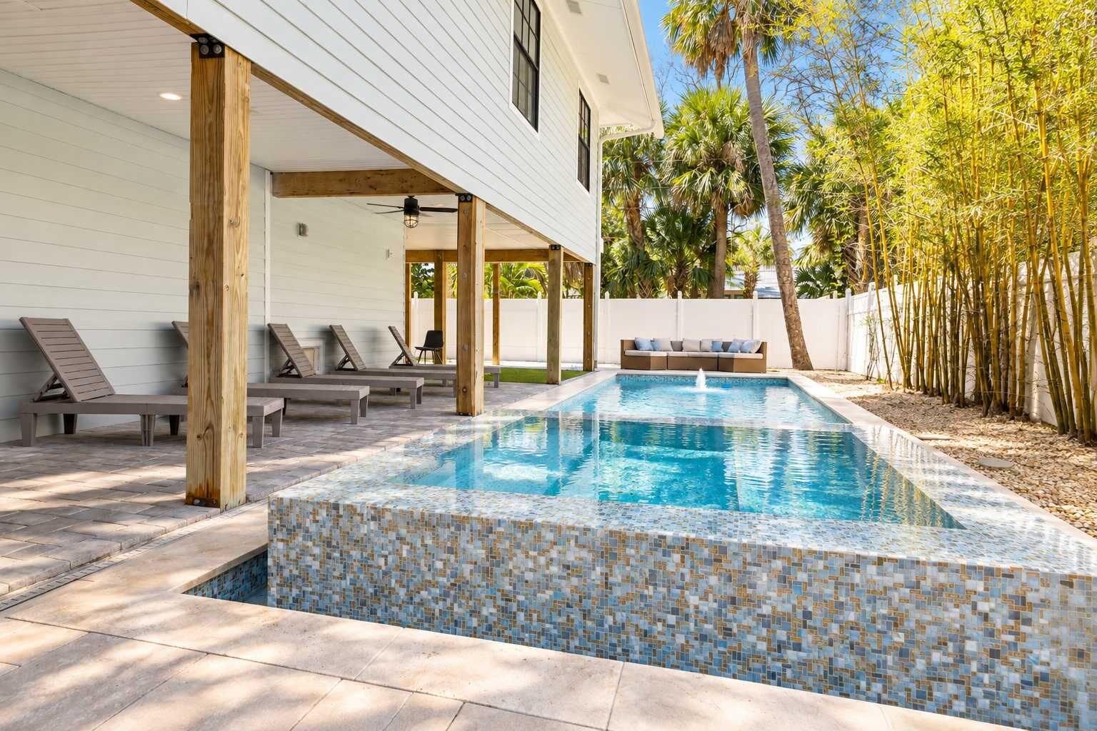 Una piscina rectangular con borde de mosaicos, rodeada de sillones de descanso y una terraza de madera.