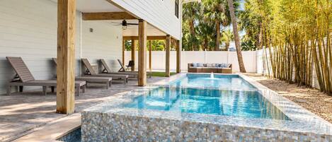 Una piscina rectangular con borde de mosaicos, rodeada de sillones de descanso y una terraza de madera.