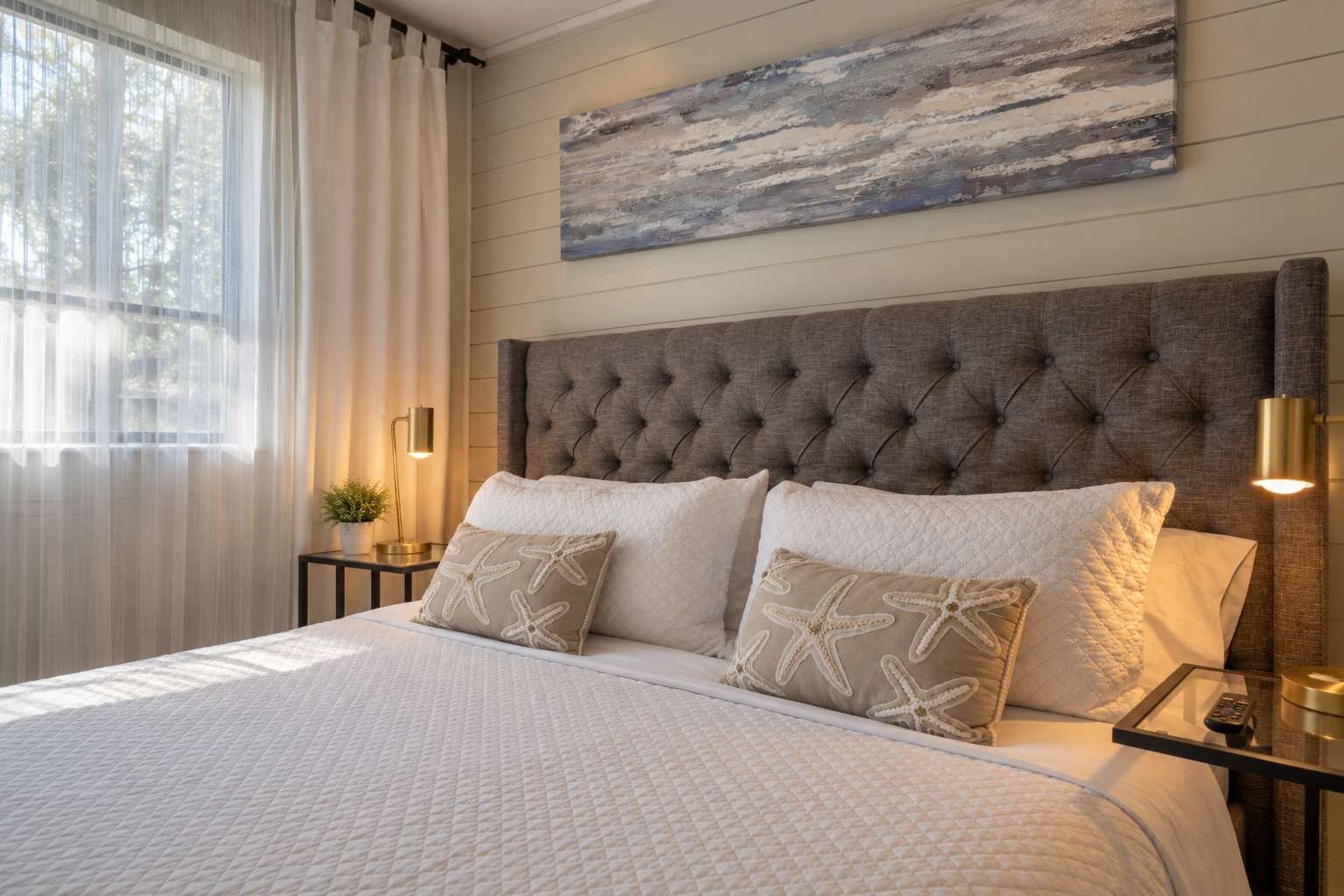 A neatly made bed with a quilted white duvet, two pillows with starfish designs, a tufted headboard, a painting above the bed, and a bedside lamp.