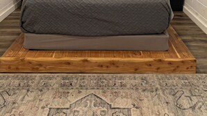Un dormitorio con techo inclinado, un ventanal grande, una cama con colcha gris, dos mesitas de noche y una alfombra estampada.