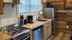 Cocina funcional con estufa a gas, lavavajillas y refrigerador. Se observa una ventana con persianas, paredes de madera y una lámpara colgante.