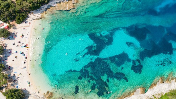 Luchtbeeld van een strand met helder turquoise water, een rotsachtige kustlijn en een zandstrand met parasols en mensen.