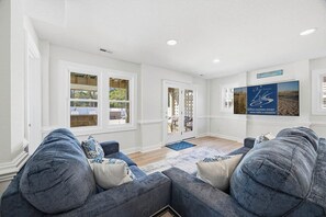 A spacious living room with a large gray sectional sofa, a blue rug, and a view of the outdoors through large windows. - Tides Rollin' In | Private Pool, Hot Tub, & Fenced in Yard (Nags Head)