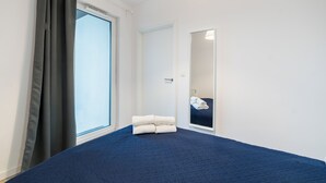 Et hotelværelse med en stor seng, et vindue med gardiner, et spejl og en pænt redt seng med håndklæder.