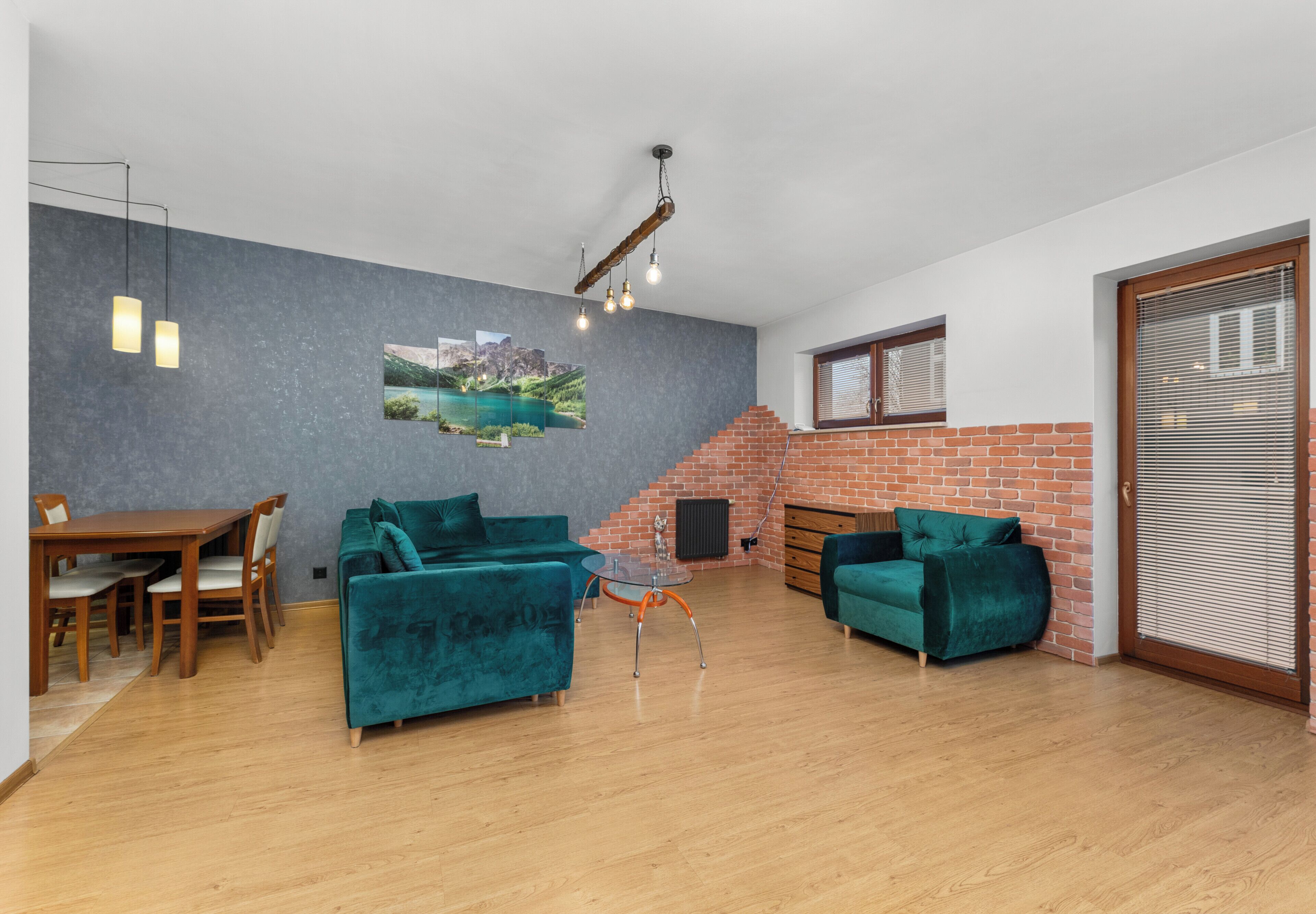 Amplia sala de estar con dos sofás verdes, una mesa de comedor de madera y una mesa de centro de vidrio. Se observa una pared de ladrillo y una escalera al fondo.