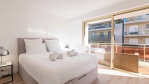 Moderni hotellihuone, jossa on suuri sänky, yöpöytä, lamppu ja parveke, jolta on näkymä kaupunkiin.