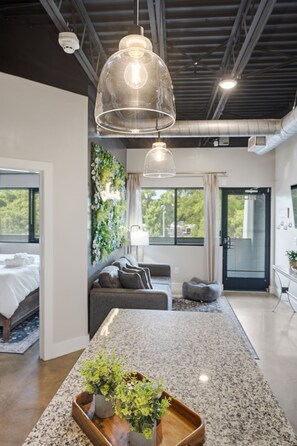 A modern hotel room with a bed, a sofa, a dining table with plants, and a large window. - 1BR Condo - King Bed - 10mins to Broadway (Nashville)