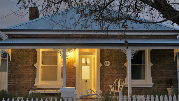 A brick house with a white picket fence, a number '104' on the wall, and a porch with a chair. - Birchfrost cottage | Quintessential cosy | Central Orange (Orange)