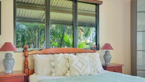 Una cama bien hecha con un edredón estampado, dos lámparas de noche y una ventana con vistas a palmeras.