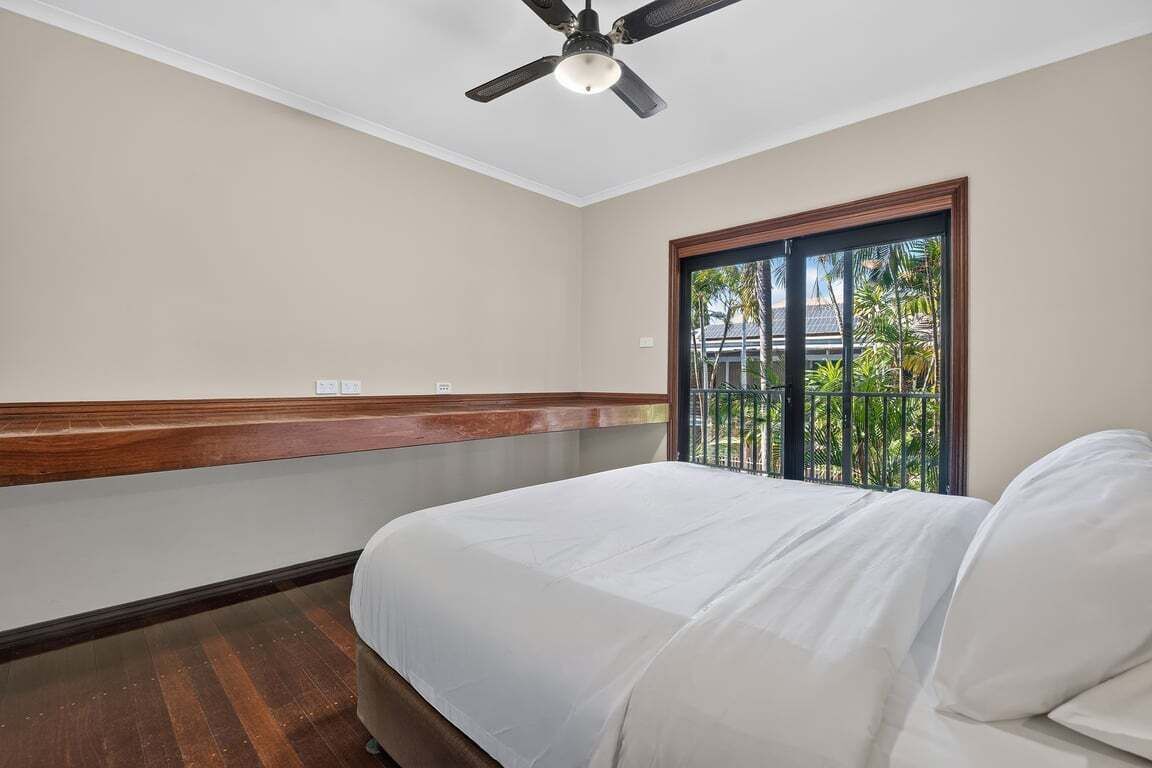 Quarto de hotel com cama, ventoinha no teto, janela com vista para palmeiras e chão de madeira.