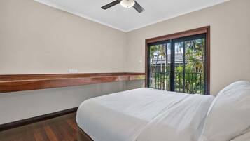 Quarto de hotel com cama, ventoinha no teto, janela com vista para palmeiras e chão de madeira.