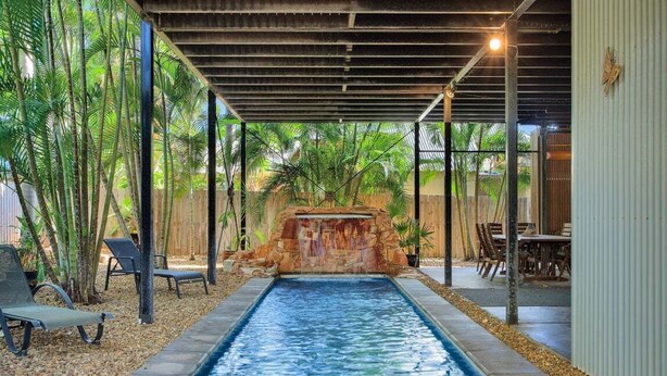 Una piscina rectangular con cascada, rodeada de palmeras y una zona de estar cubierta.