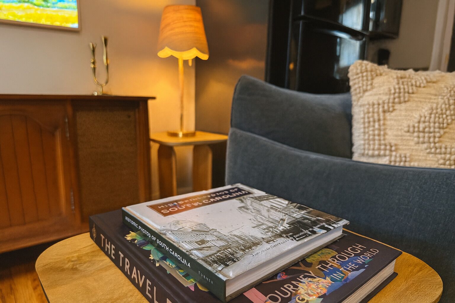 A stack of books on a small wooden table in a cozy living room with a sofa and a lamp in the background.