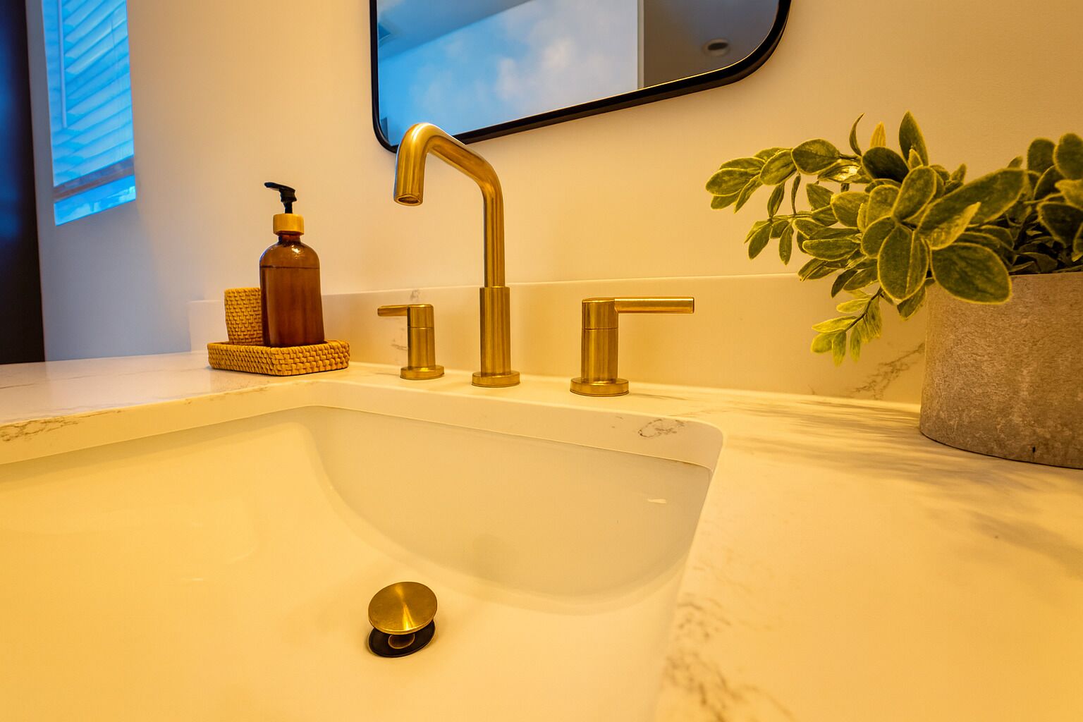 Un baño con un lavabo blanco, grifos dorados, un dispensador de jabón, una planta en maceta y un espejo en la pared.