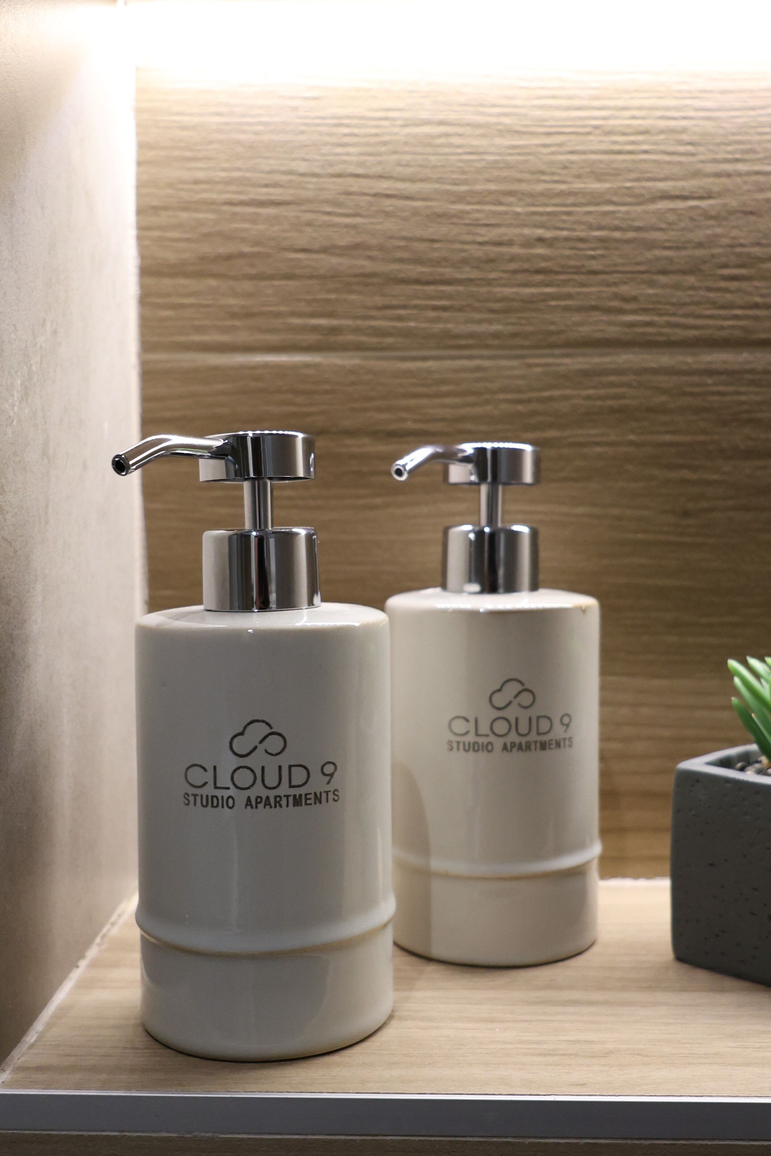 Two white pump dispensers with the text "CLOUD 9 STUDIO APARTMENTS" on a wooden shelf.
