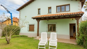Una casa de dos pisos con techo de tejas, un porche cubierto y un pequeño jardín con dos sillas blancas.