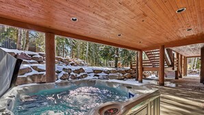 Un jacuzzi en un espacio exterior cubierto con vigas de madera, rodeado de rocas y árboles cubiertos de nieve.