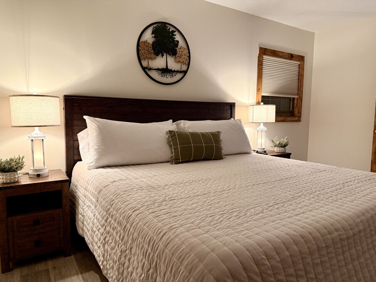 A neatly made bed with a quilted cover, two bedside lamps, a wall-mounted tree artwork, and a window with blinds.