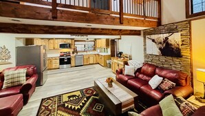A spacious living area with a high ceiling, wooden beams, a kitchen in the background, and a stone accent wall. - Yough Lake Streamside Cottage w/ Hot Tub, Pavilion, Firepit (Confluence)