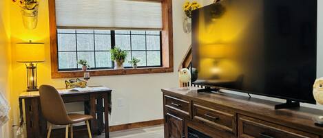 A living room with a large flat-screen TV, wooden furniture, a fireplace, and a window with blinds.
