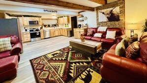 A spacious living room with a red leather sofa set, a patterned rug, and a kitchen in the background. - Yough Lake Streamside Cottage w/ Hot Tub, Pavilion, Firepit (Confluence)