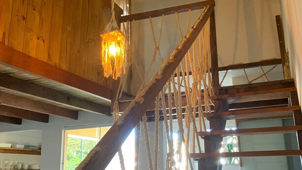 Uma escada de madeira com corrimão de corda, luminária pendente e vista para a área da cozinha com prateleiras e uma janela.