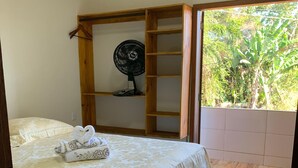 Quarto com cama, guarda-roupa de madeira, ventoinha e vista para o verde através de uma janela.