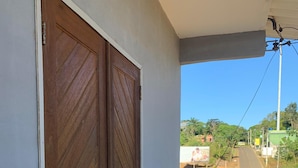 Varanda com porta de madeira, chão de azulejos e vista para a rua com vegetação e edifícios.