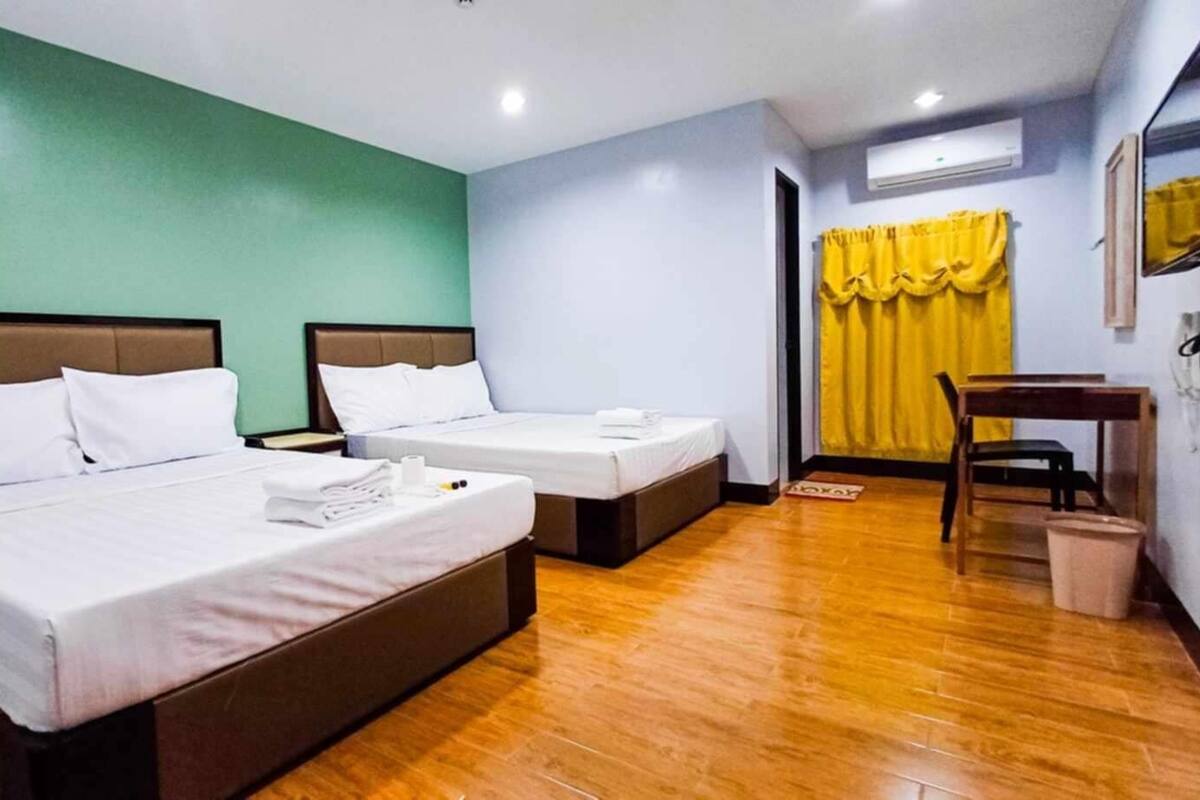 A hotel room with two beds, wooden flooring, a small table, a chair, and a yellow curtain.