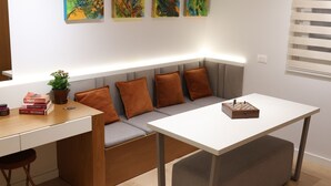 Una habitación moderna con un escritorio blanco, un sofá gris con cojines naranja y un tablero de ajedrez en el escritorio. Hay tres cuadros abstractos en la pared y una planta en maceta sobre el escritorio.