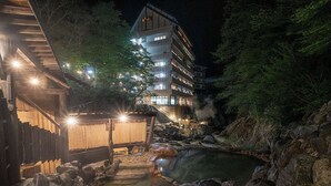 A traditional Japanese onsen with a wooden bathhouse, a stone hot spring, and a multi-story building in the background. - Livemax Resort Kawamata Onsen (Nikko)