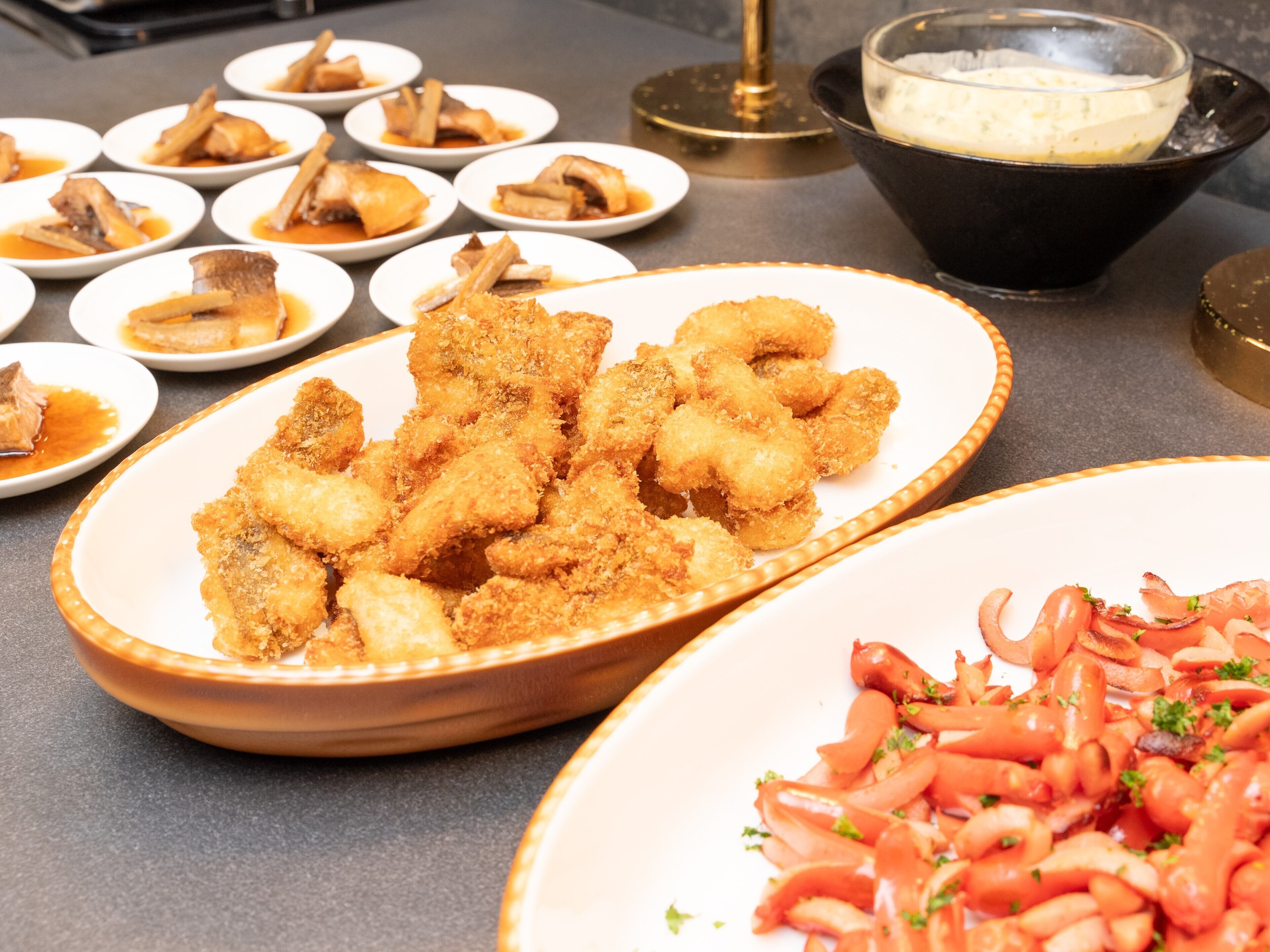 A buffet table with various dishes including breaded chicken wings, a bowl of soup, and a plate of sliced tomatoes.
