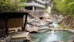 A hot spring area with a wooden structure, a stone pool, and a building in the background. - Livemax Resort Kawamata Onsen (Nikko)